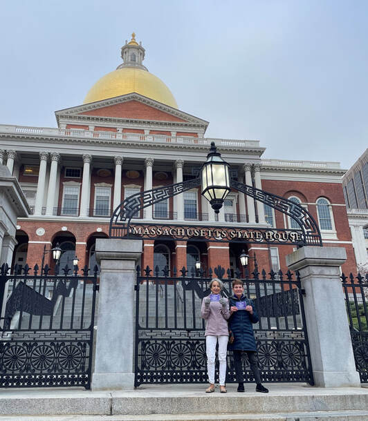 Two BCCR members stand in front of the MA State House holding flyers that say, 